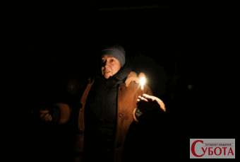 Жінка у теплому зимовому одязі та синій шапці стоїть у темному приміщенні, її обличчя яскраво освітлене полум'ям єдиної свічки